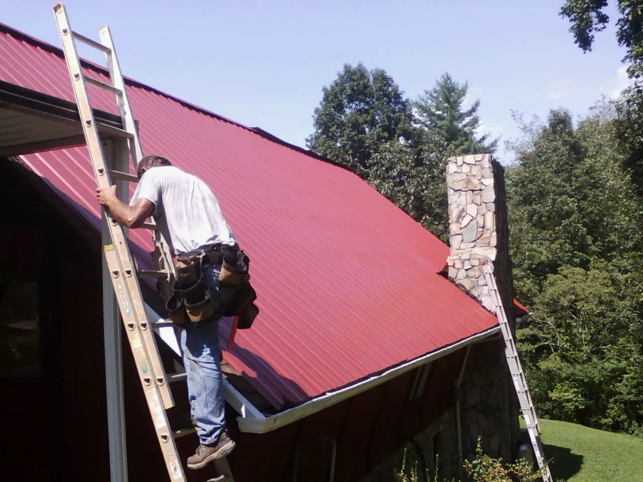Roofer on a ladder beside a red metal roof and stone chimney — Soffit & Fascia Repair by Roofing Brothers in Narvon